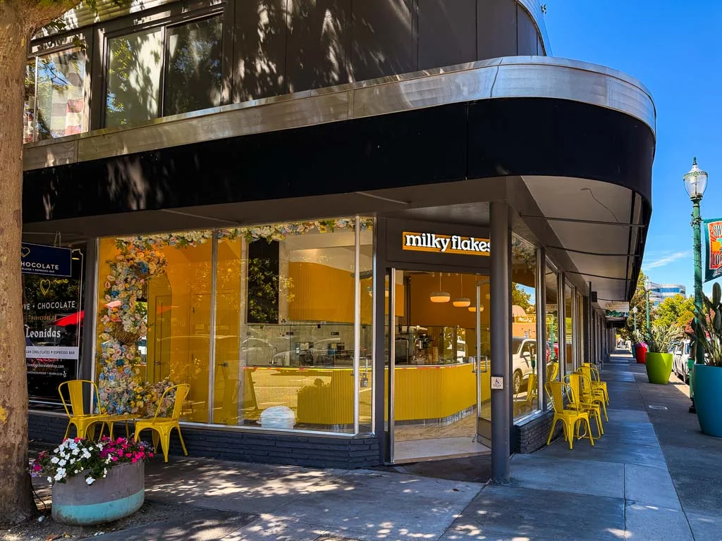 The Milky Flakes corner storefront on a sunny day, with the glowing milky flakes sign over the door, yellow walls inside and yellow chairs on the sidewalk.